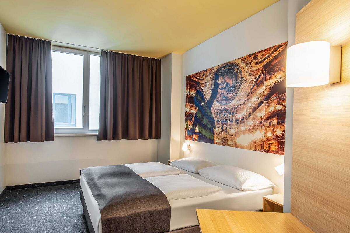 A hotel room with a bed, a nightstand, a desk, a window with curtains, and a large tapestry on the wall.