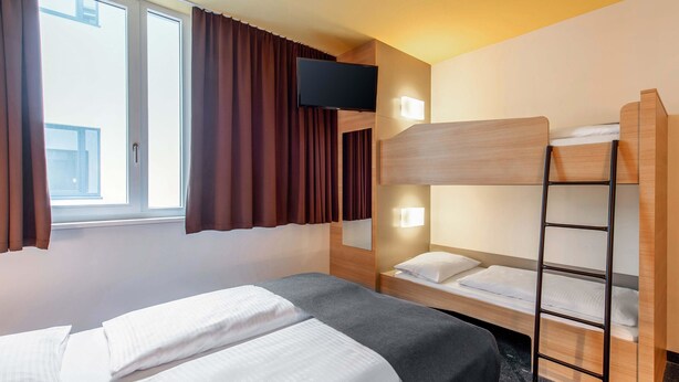 Habitación de hotel con literas, una cama individual, un televisor de pantalla plana montado en la pared y una ventana con cortinas.