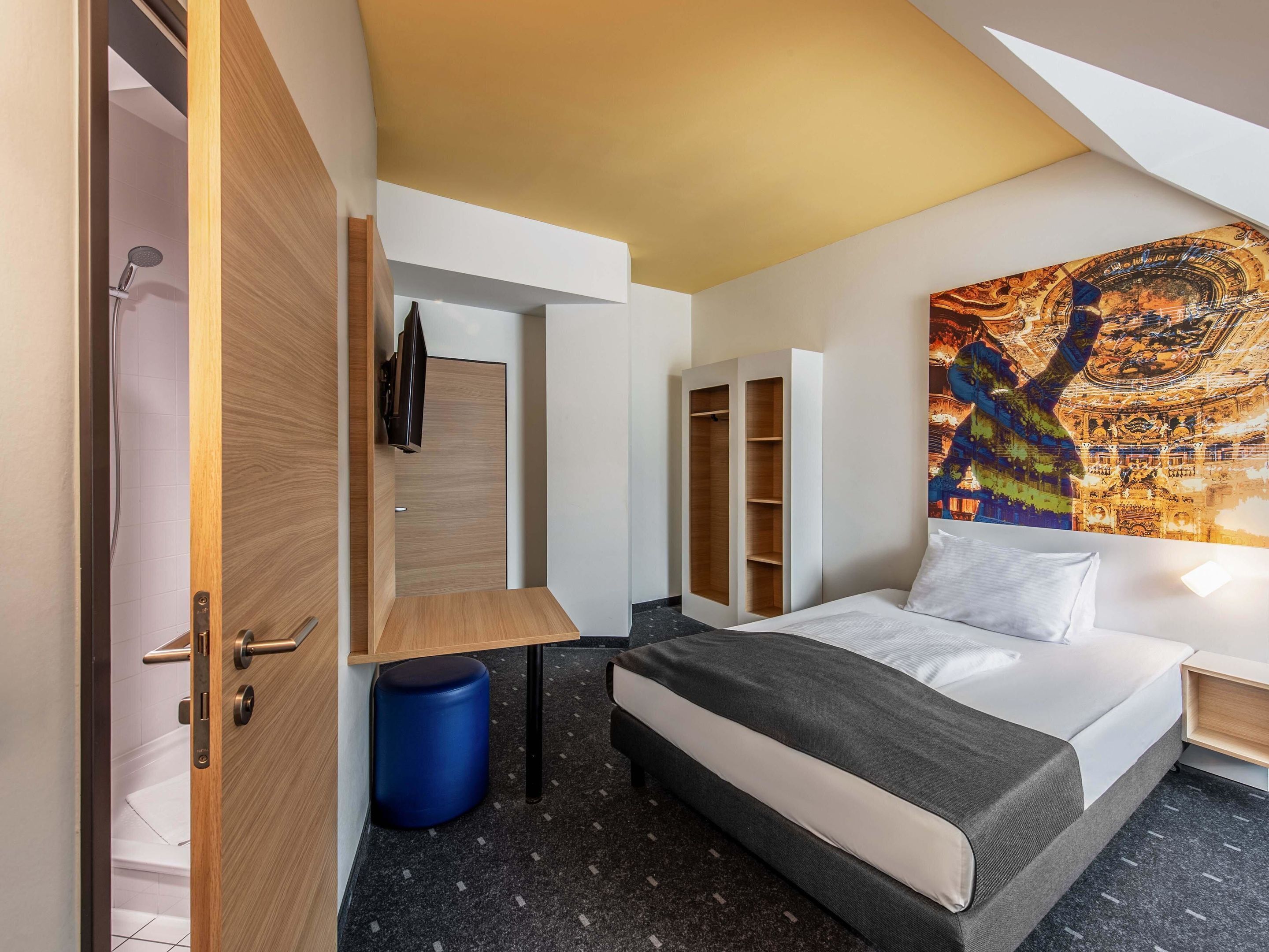 A modern hotel room with a bed, a desk, a wardrobe, and a large painting on the wall.