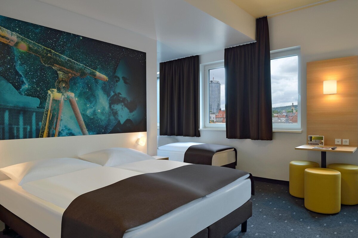 Una habitación de hotel con una cama grande, un escritorio con silla, una mesita de noche con lámpara y un ventanal con cortinas.