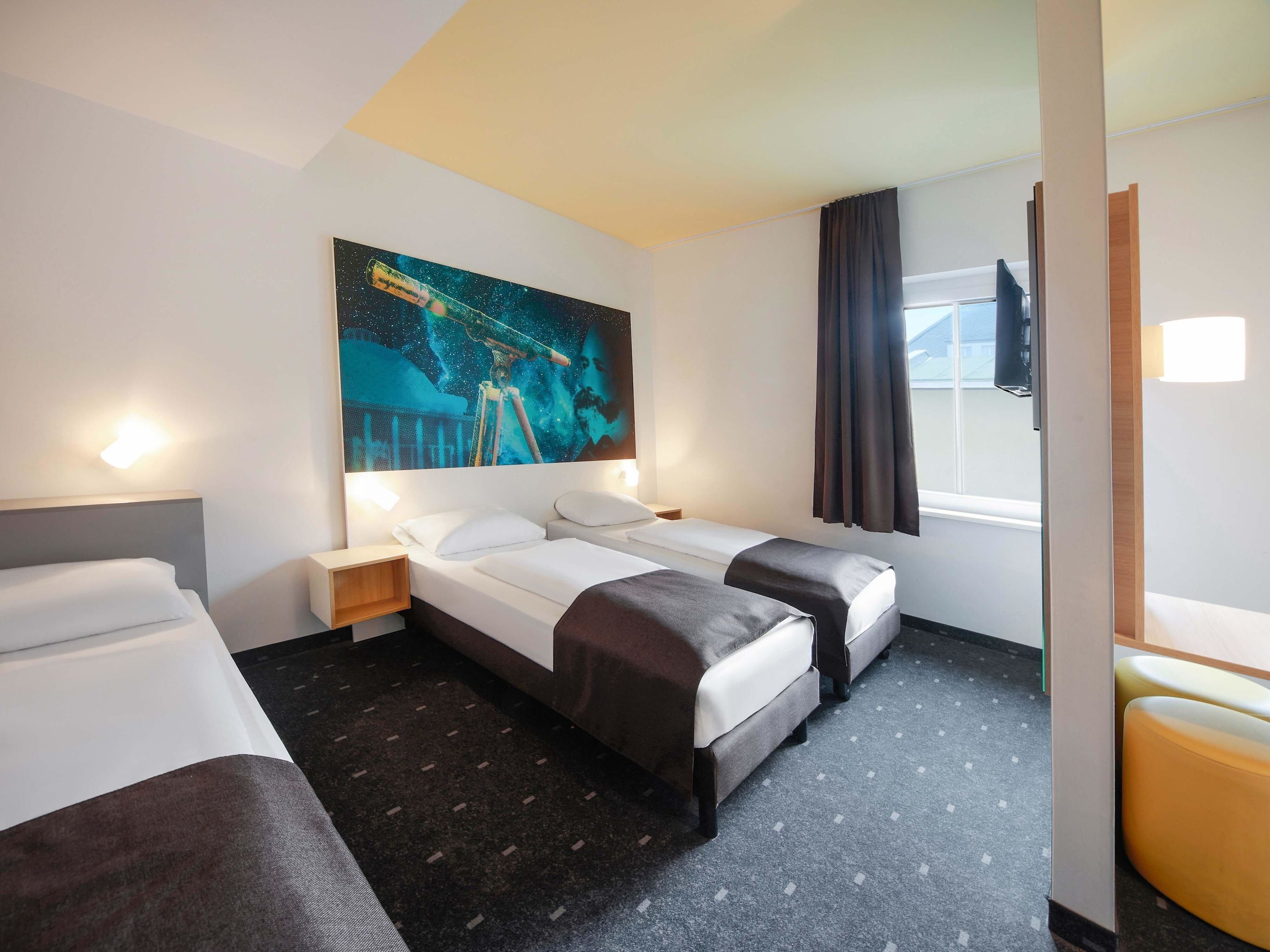 Quarto de hotel com duas camas, um grande mural na parede, uma janela com cortinas e um pequeno espaço para sentar.