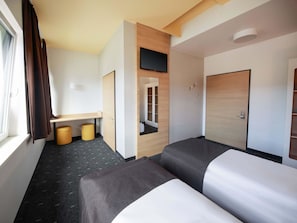 Habitación de hotel con dos camas, un escritorio, una silla, un taburete pequeño, un espejo y un armario.
