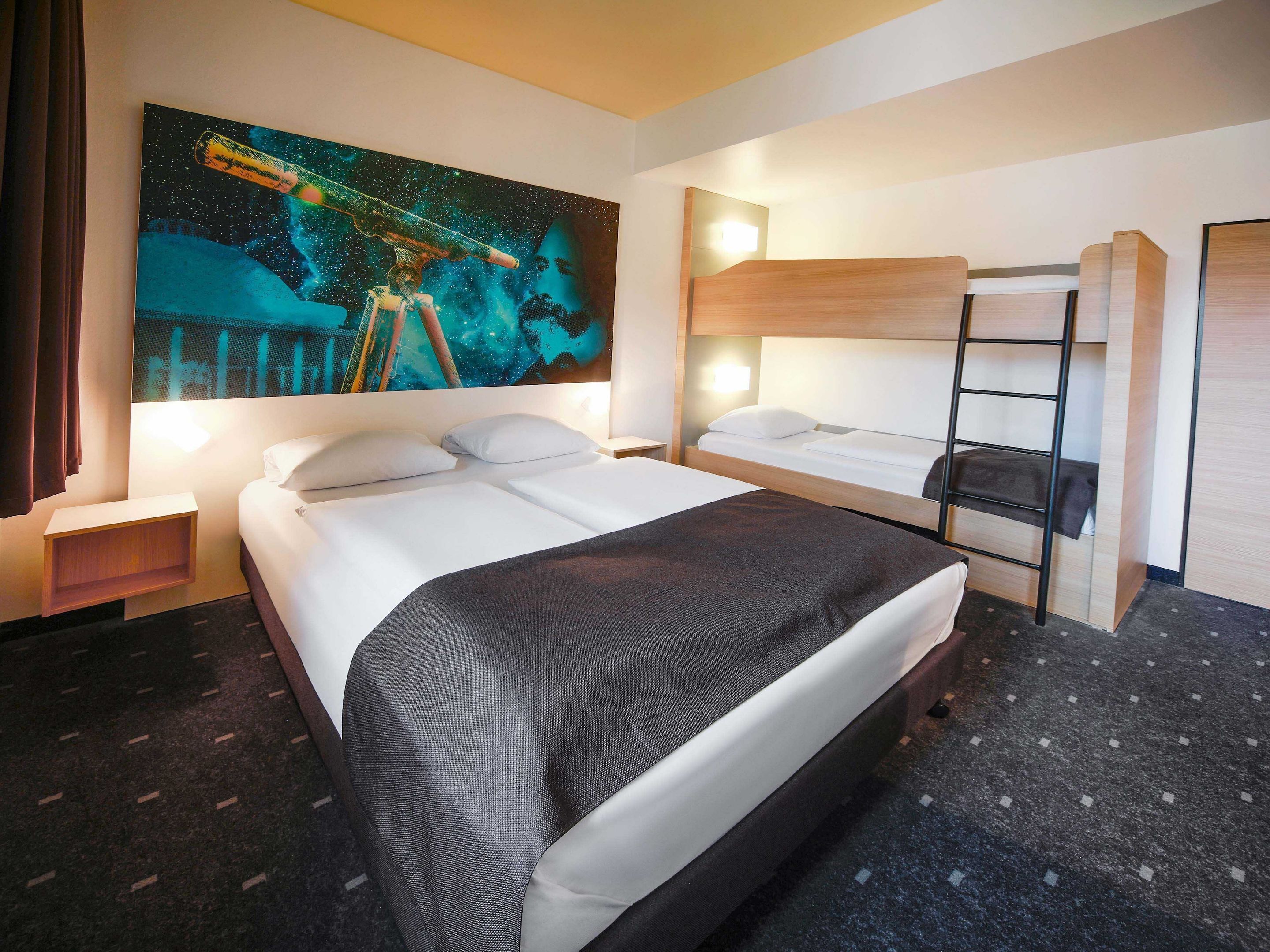 Ein Hotelzimmer mit einem großen Bett, einem kleineren Bett, einem Etagenbett, einem Nachttisch, einer Lampe und einem Bild mit Weltraumdarstellung.