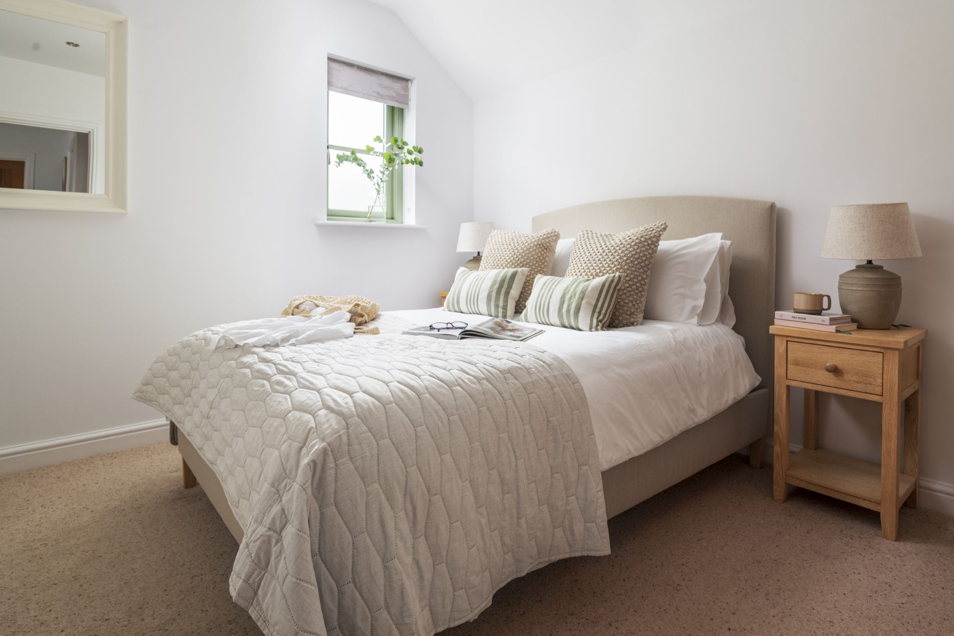A bedroom with a bed, a nightstand, a lamp, a window with a view of greenery, and a mirror on the wall.
