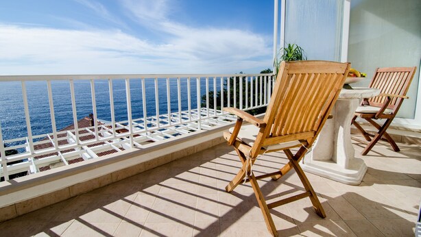 A balcony with a white railing, wooden chairs, a potted plant, and a view of the ocean.