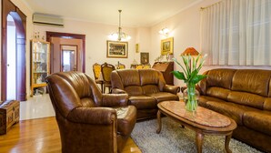 A living room with leather sofas, a coffee table, and a vase with plants. There is a chandelier, framed pictures on the wall, and a door leading to another room.