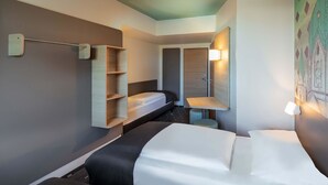 Una habitación de hotel con cama, escritorio, silla, armario y estante en la pared.