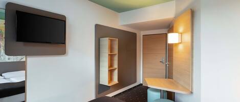 A hotel room with a bed, a TV mounted on the wall, a wooden shelf, a small stool, and a door with a light.