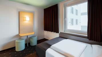 Ein Hotelzimmer mit Bett, Nachttisch, Lampe, einem Fenster mit Vorhängen und Blick auf Gebäude.