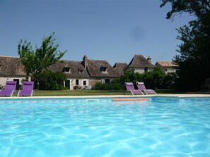 A swimming pool with clear blue water, surrounded by lounge chairs and a building with a tiled roof.
