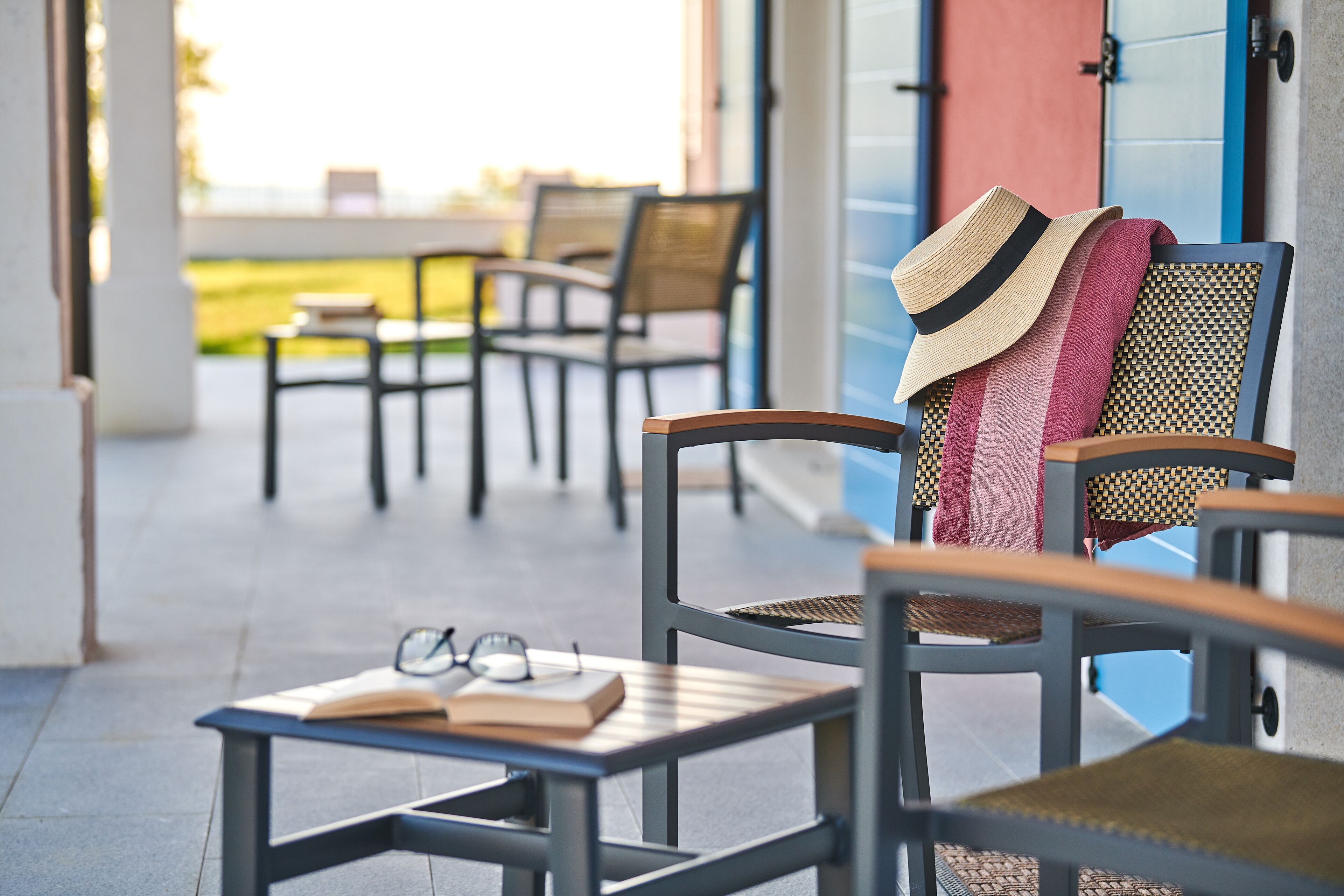 A chair with a straw hat and a book on a table, another chair with a towel, and a view of the outdoors.