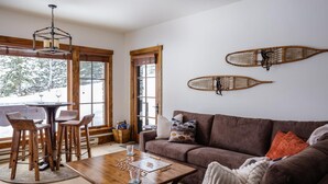 A cozy living room with a brown sofa, wooden coffee table, and two bar stools. There is a window with a view of trees and a wooden ceiling. - Hot Tub| Ski-In/ Ski-Out | Sonos Sound (Big Sky)