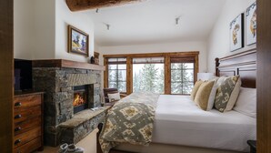 A bedroom with a stone fireplace, wooden beams, a large bed with patterned bedding, and a view of trees through the windows. - Hot Tub| Ski-In/ Ski-Out | Sonos Sound (Big Sky)
