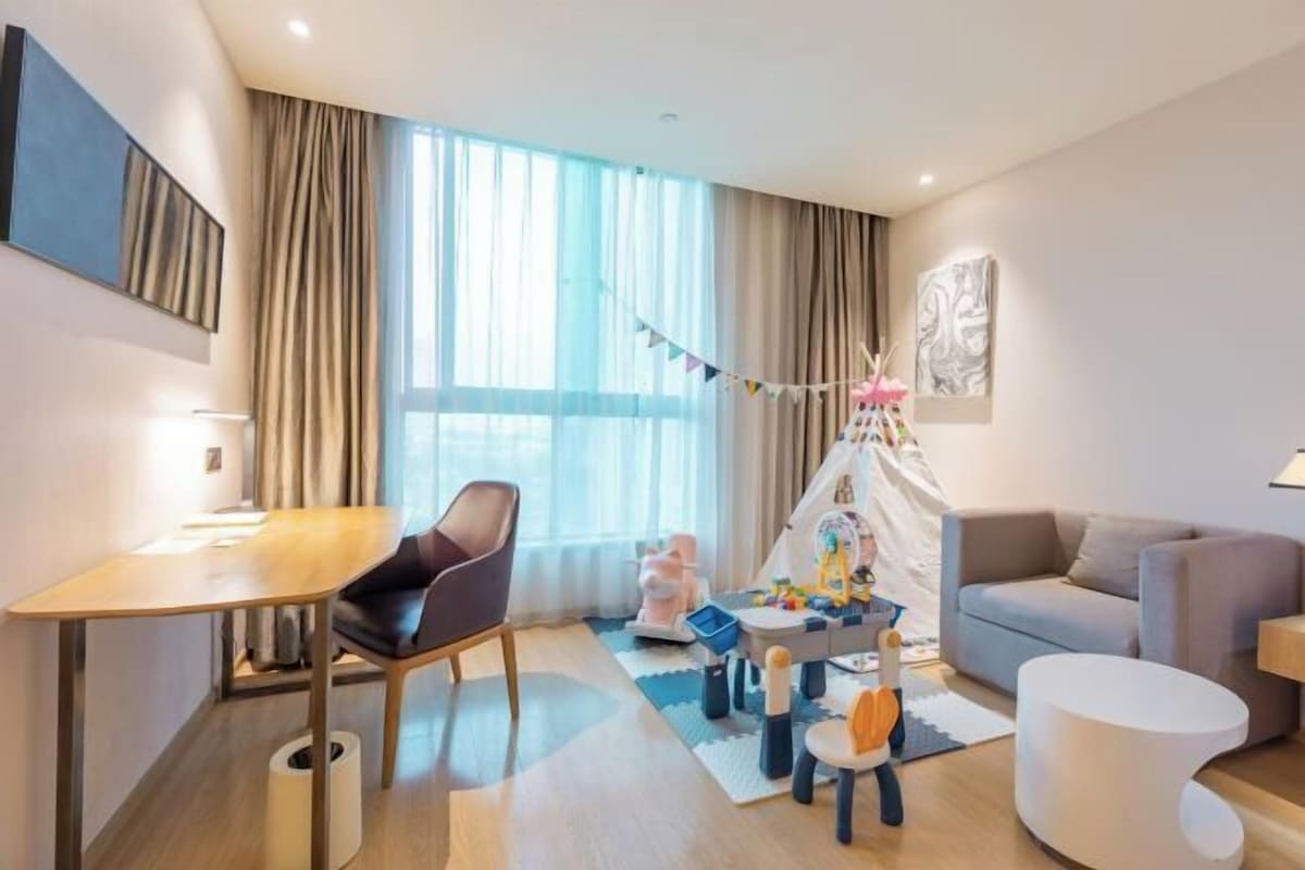 A modern living room with a wooden table, a grey sofa, a white ottoman, and a play area with toys and a small tent.
