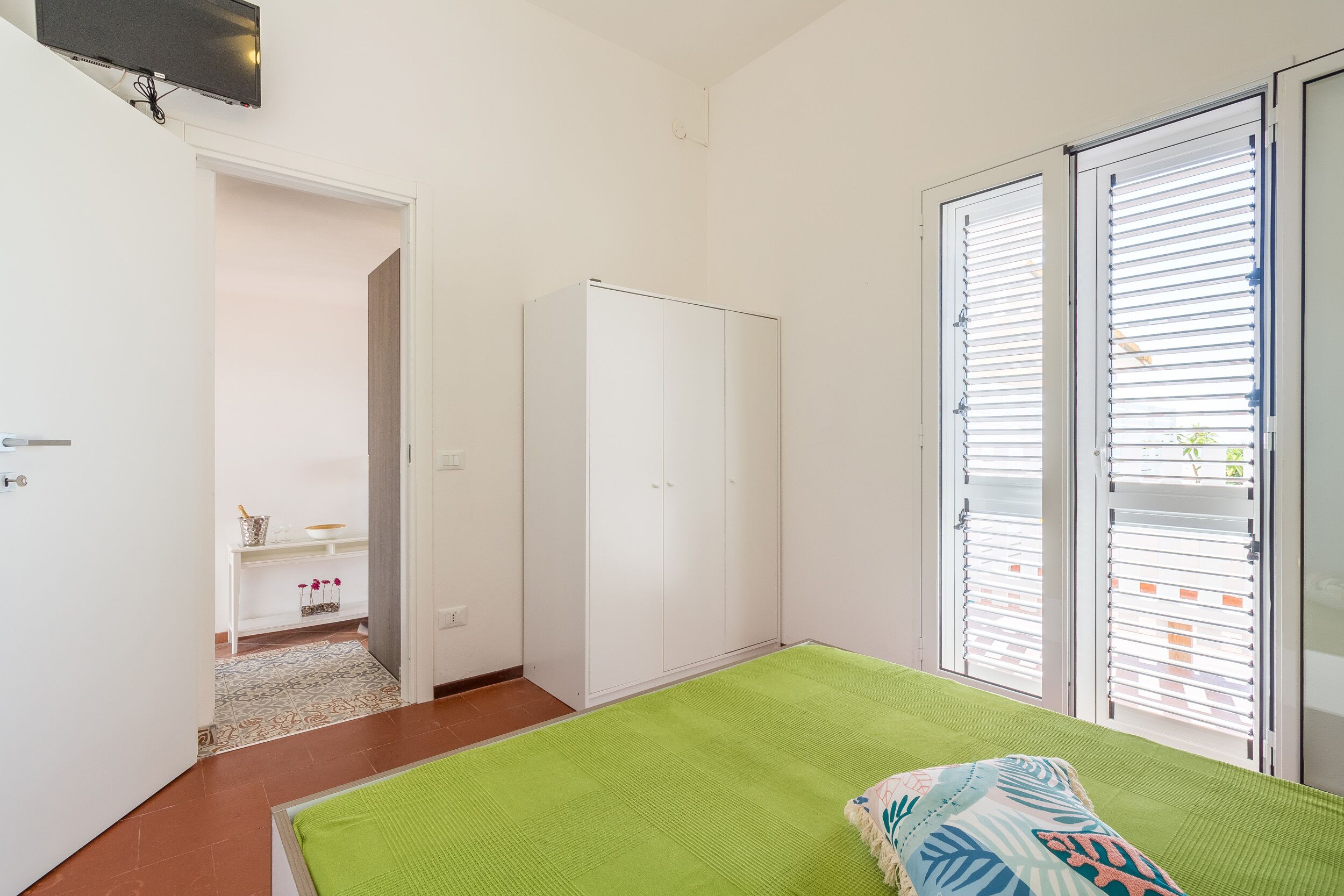 Une chambre avec un tapis vert, une armoire blanche et une porte-fenêtre coulissante avec des stores.