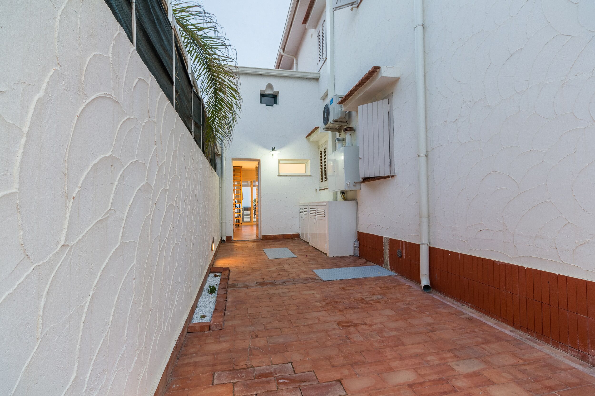 A narrow, tiled corridor with a white textured wall on one side and a wooden baseboard on the other. A door leads to an interior space.