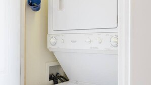 A compact laundry room with a front-loading washer and dryer, a blue bucket on the wall, and a small shelf with items. - DT~ Free parking~ Sunset river views~ WiFi~ W/D (Wilmington)