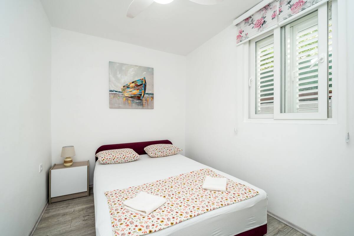 A neatly arranged bedroom with a bed, a bedside table with a lamp, a painting on the wall, and a window with blinds.