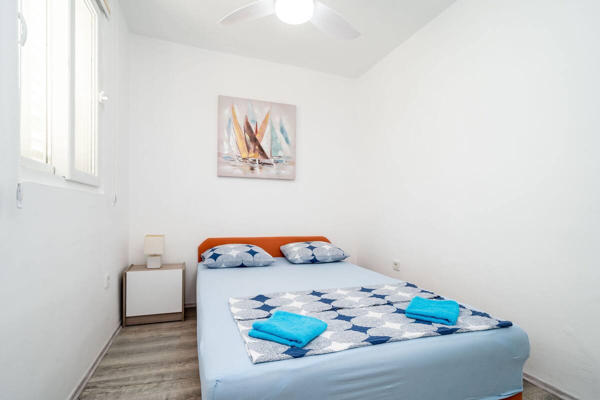 A bedroom with a bed, a nightstand, a lamp, a ceiling fan, and a painting on the wall.
