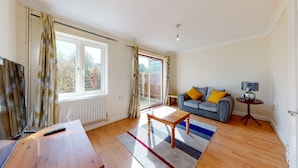 A spacious living room with a wooden floor, a sofa with yellow cushions, a coffee table, and a lamp on a side table. - Amazing 2 Bed House w/ Private Parking & Private Garden (London)