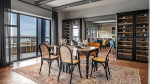 A spacious dining area with a large table and chairs, a view of the ocean through large windows, and a wine cellar with shelves of bottles. - Belle Maroc (Cape Town)