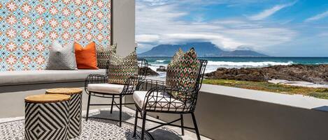 A balcony with a patterned wall, two chairs with cushions, a round table, and a view of the ocean and a mountain.