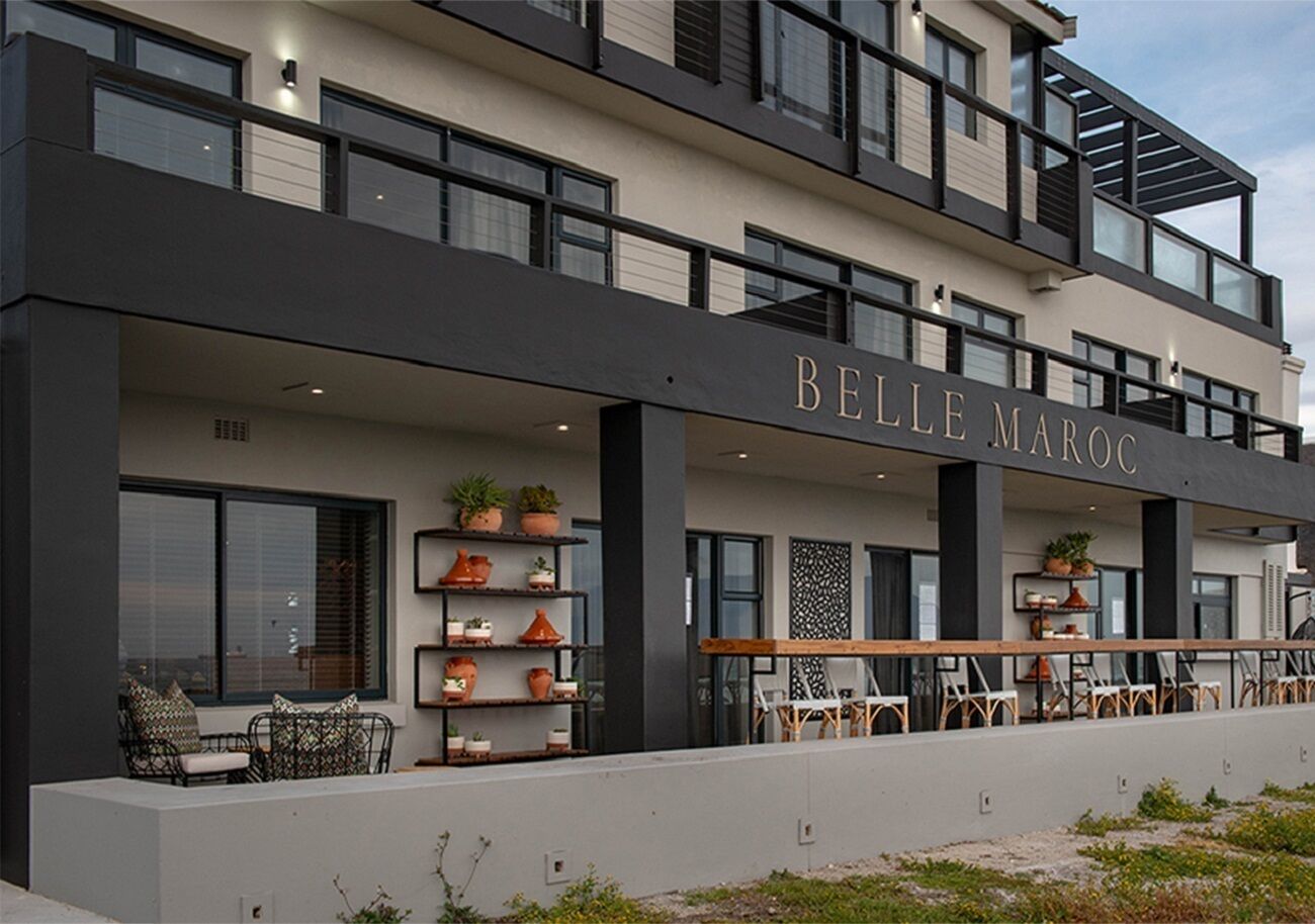 Un bâtiment moderne dont la façade porte l’inscription « BELLE MAROC ». On y trouve des espaces de détente en plein air avec des chaises et des tables, et des plantes en pot sont disposées sur des étagères.