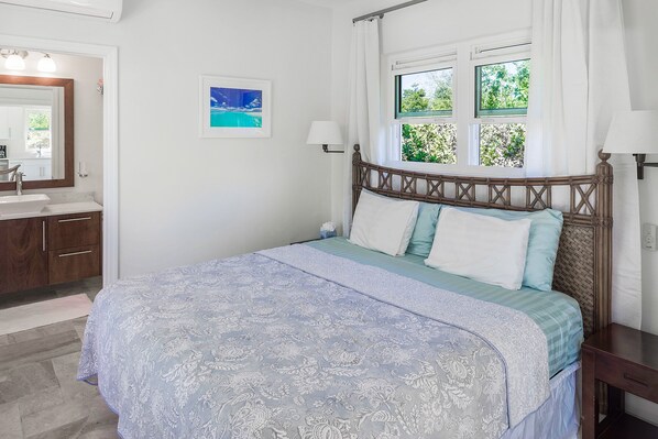 A bedroom with a bed, a window with curtains, a bedside table, and a bathroom with a mirror and sink.