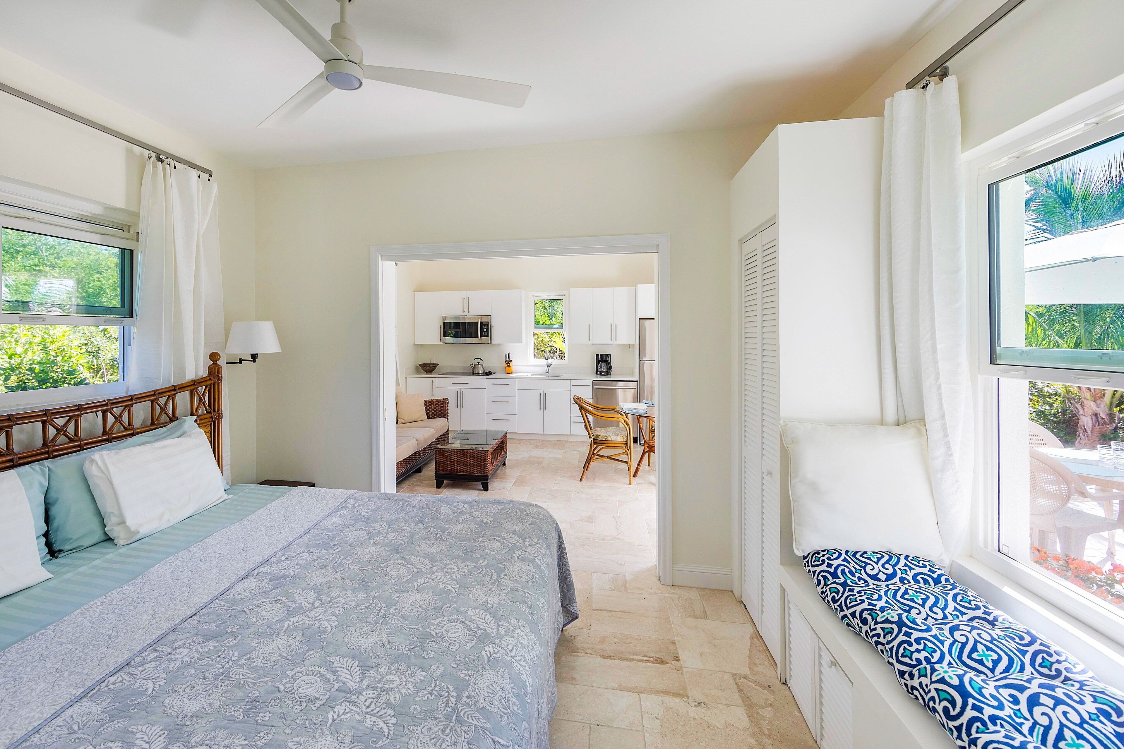 A bedroom with a bed, a window seat, and a view of the kitchen and dining area.