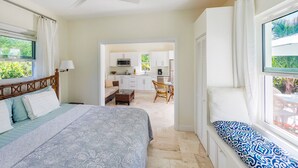 A bedroom with a bed, a window seat, and a view of the kitchen and dining area.