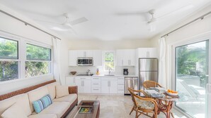 A modern kitchen with white cabinets, a dining area with wicker furniture, and large windows offering a view of greenery.