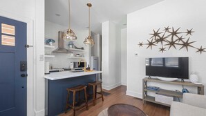 A modern kitchen with a central island, wooden stools, pendant lighting, and a TV on a wooden stand.