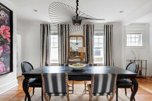 A dining room with a dark wooden table, black and white patterned chairs, a large floral painting on the left, and a unique black chandelier.