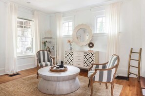 Un salon bien éclairé, avec une table basse ronde, deux fauteuils à rayures, un meuble blanc orné d’objets décoratifs et un grand miroir au mur.