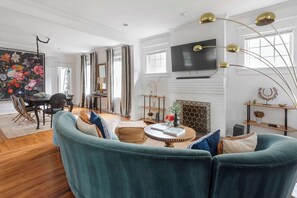 A spacious living room with a teal sofa, a fireplace, a TV mounted on the wall, and a dining area with a table and chairs. - Edgy Overlook (Atlanta)