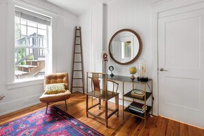 Una habitación con suelo de madera, una silla con cojín estampado, un escritorio con un globo terráqueo y libros, un espejo con marco decorativo, y una ventana que da a una escalera.