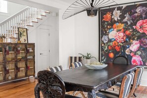 Un coin repas avec une table en bois foncé, des chaises à motifs, une fresque florale et un escalier avec des rampes blanches.