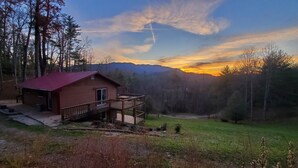 A cabin with a red roof and a wooden deck, surrounded by a forest and mountains in the background. - Majestic Peak - Mountain Views and Lake Access (Butler)