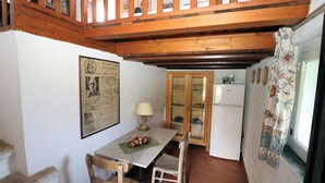 Une petite cuisine avec un plafond en bois, une table avec des chaises, un réfrigérateur et une fenêtre ornée d’un rideau à motifs floraux.
