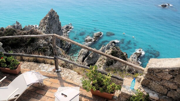 Une terrasse dotée d’un mur en pierre, d’une balustrade en bois et de plantes en pot, qui offre une vue sur une mer turquoise parsemée de rochers.
