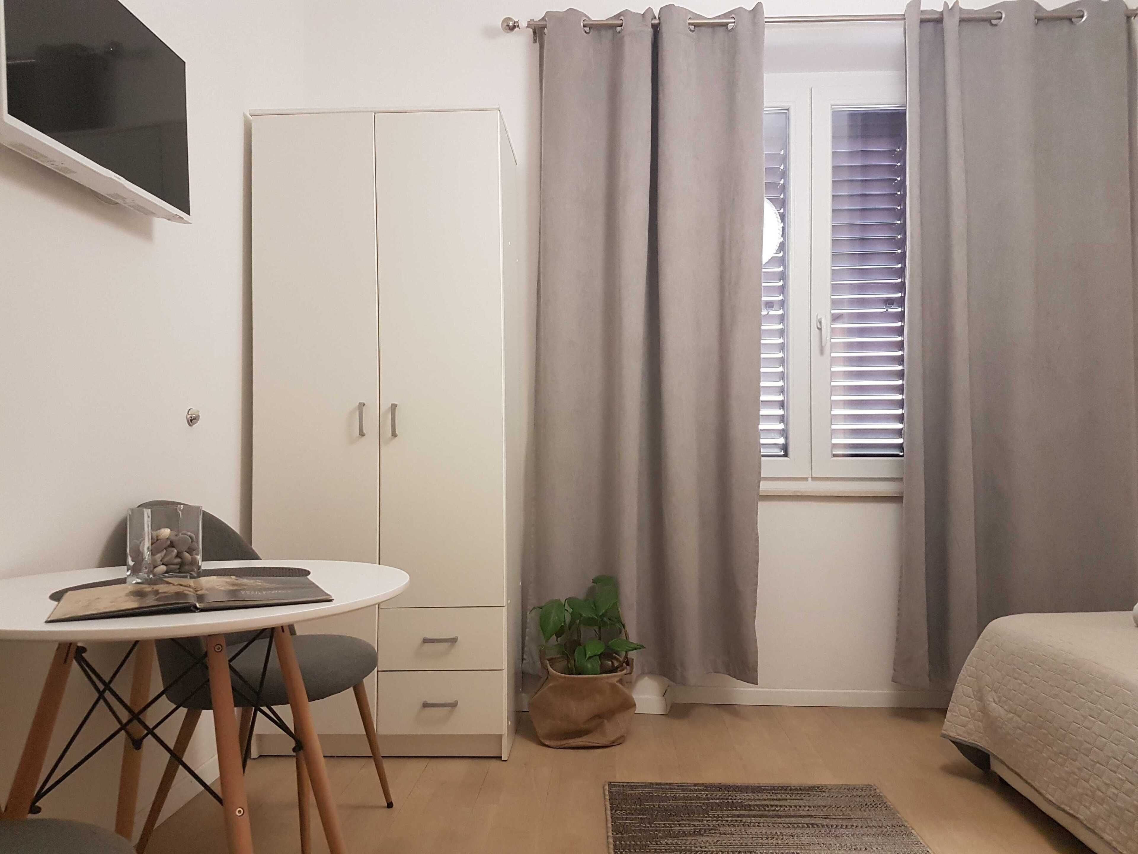 A bedroom with a bed, a desk with a chair, a wardrobe, a window with curtains, and a potted plant.