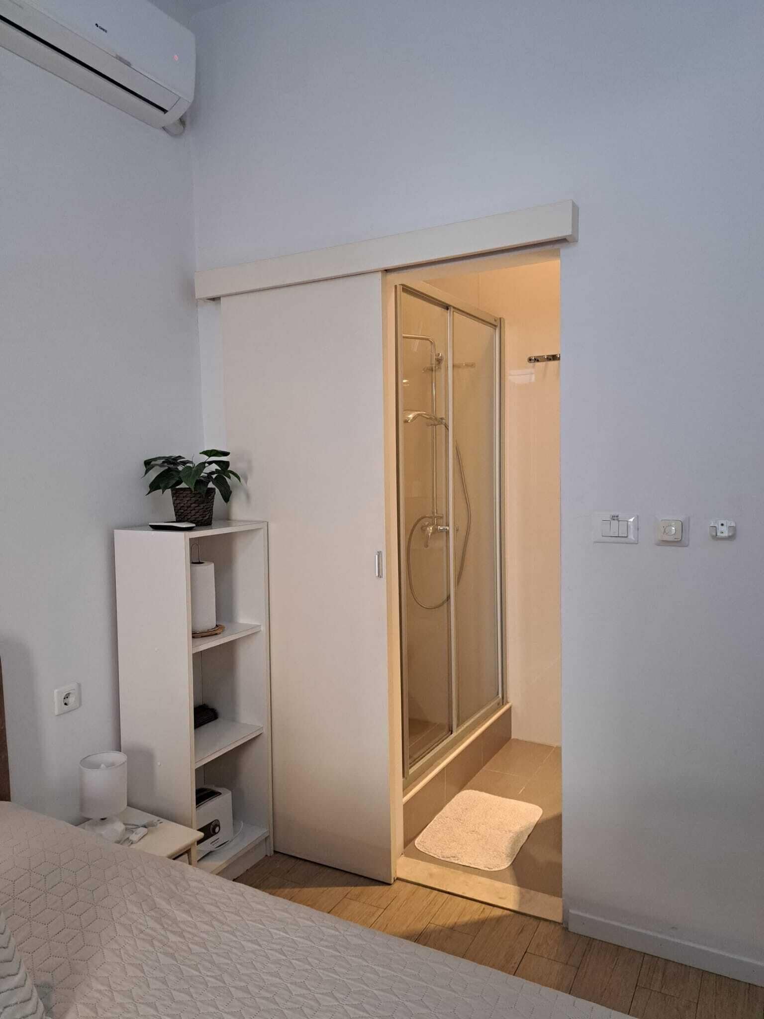 A bedroom with a bed, a white wardrobe, a shelf with a plant, and a glass-enclosed shower area.