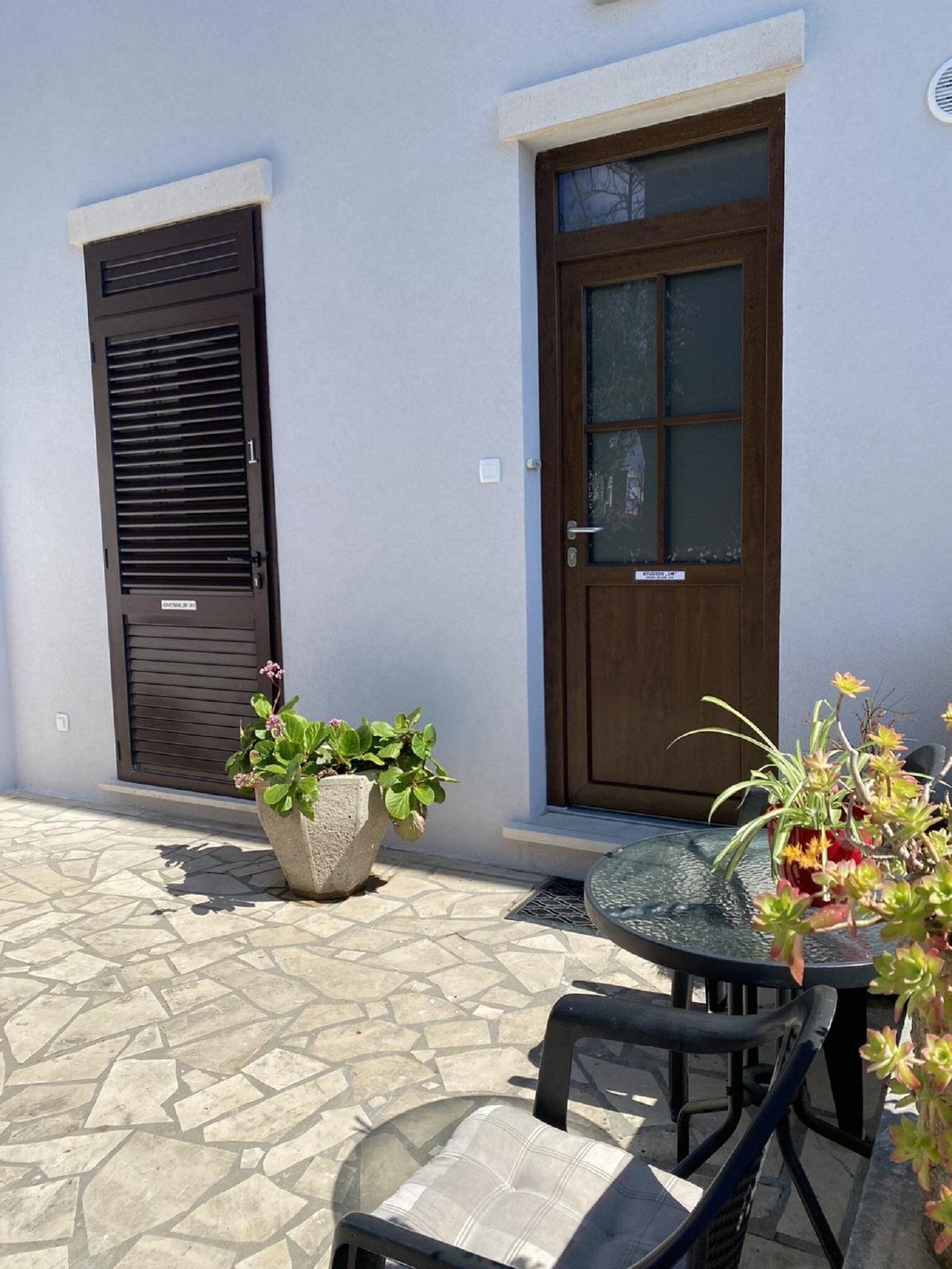 A patio with a stone floor, a glass table, and two chairs. There is a potted plant and a door with a window.