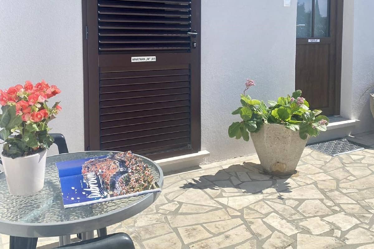 Un patio con una mesa de vidrio, dos plantas en macetas, una puerta con ventanal y un pequeño letrero en la puerta.