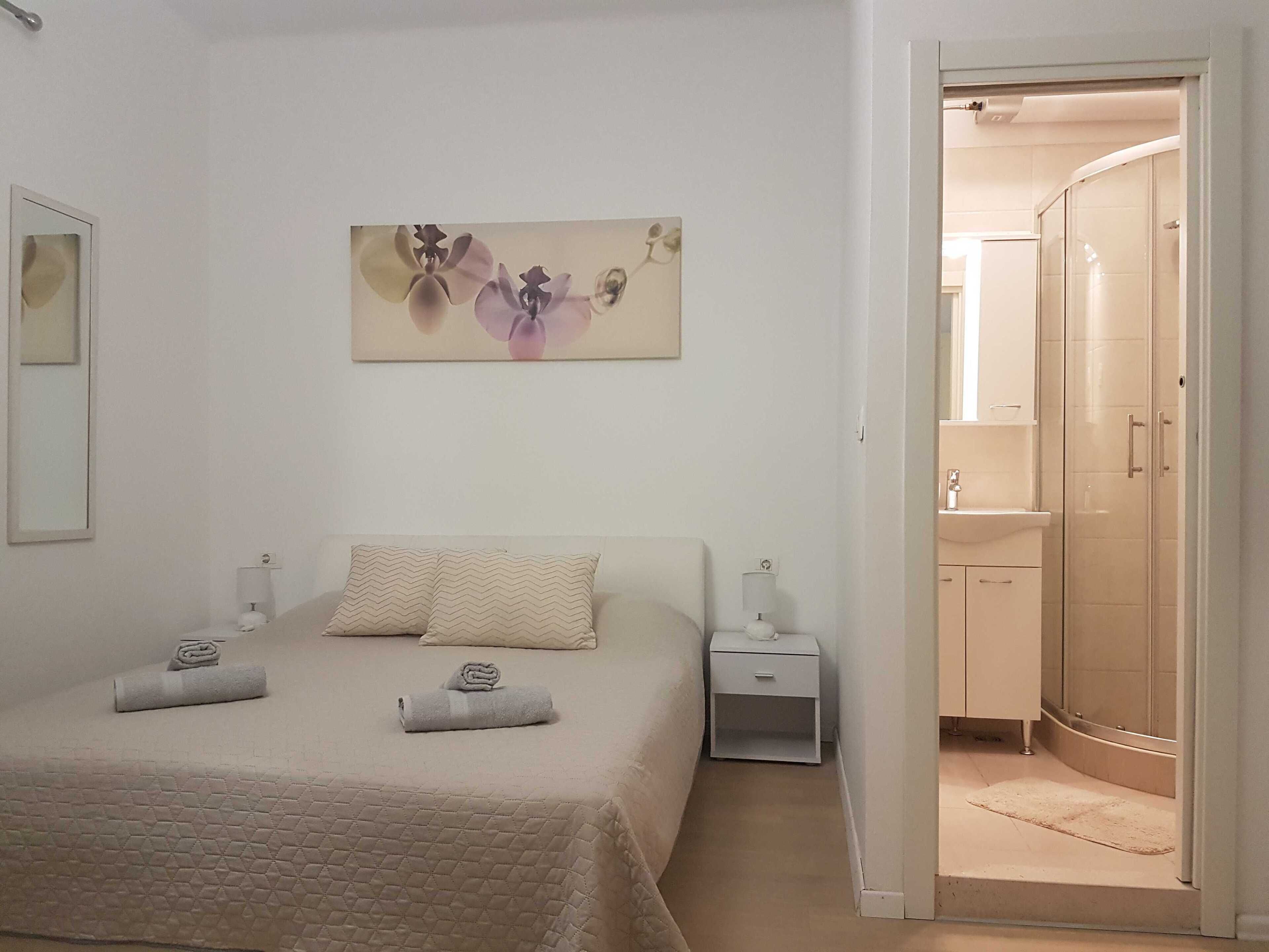 A bedroom with a bed, a nightstand, a painting, and a bathroom with a shower and sink.