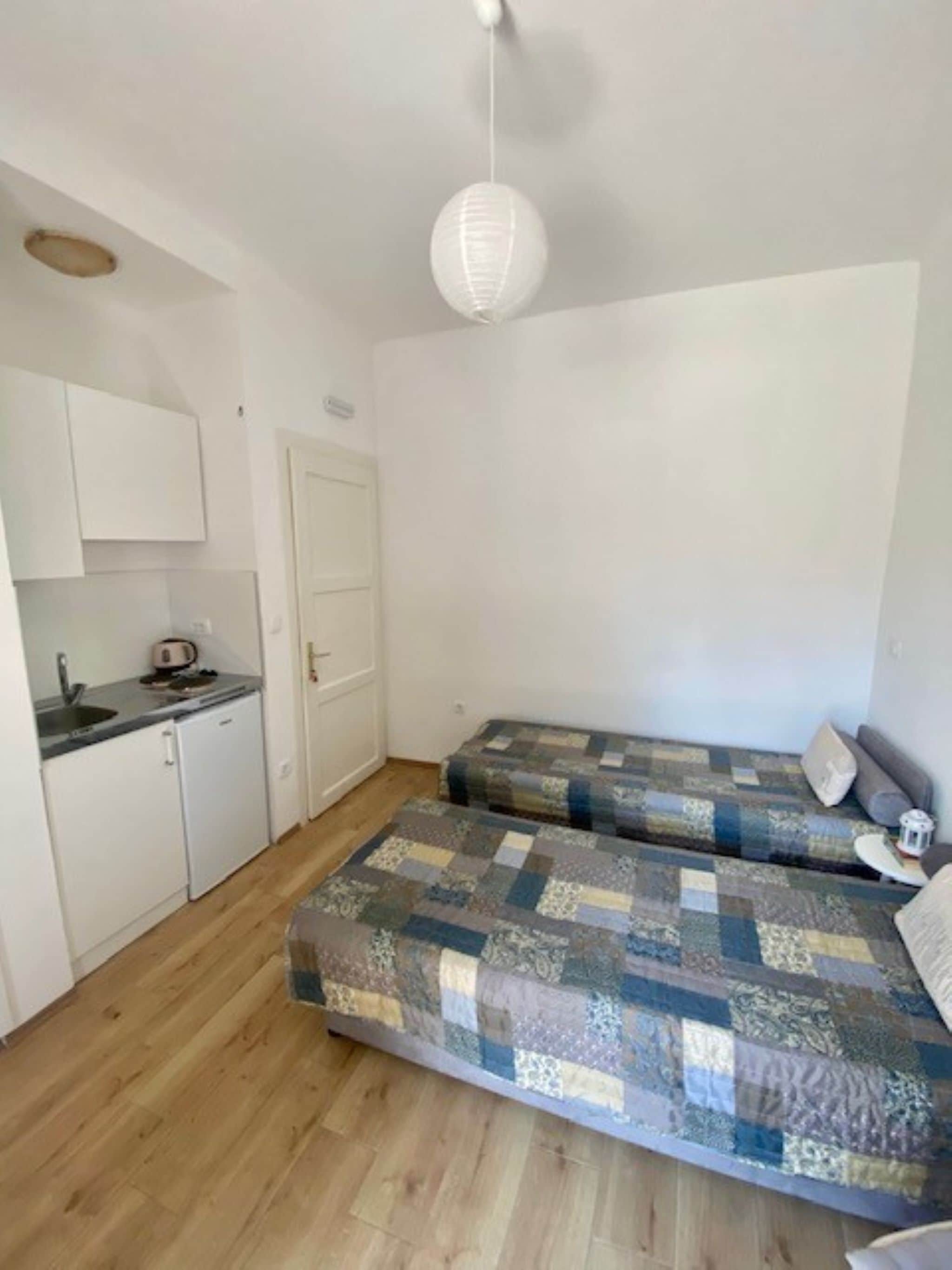 Habitación compacta con kitchenette, dos camas con ropa de cama estampada, piso de madera y lámpara colgante.