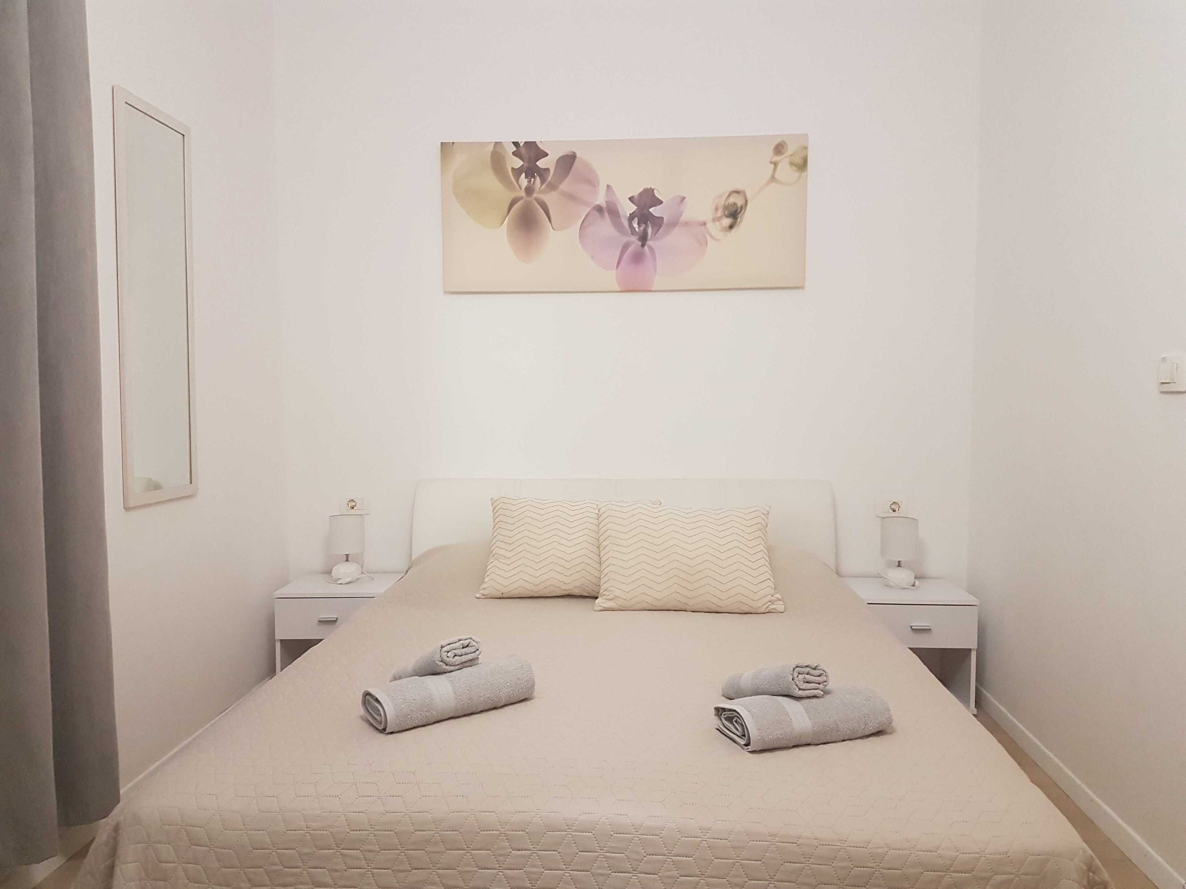 Ein ordentlich bezogenes Bett mit zwei aufgerollten Handtüchern, einem an der Wand hängenden Blumenbild und zwei Nachttischen mit Lampen.