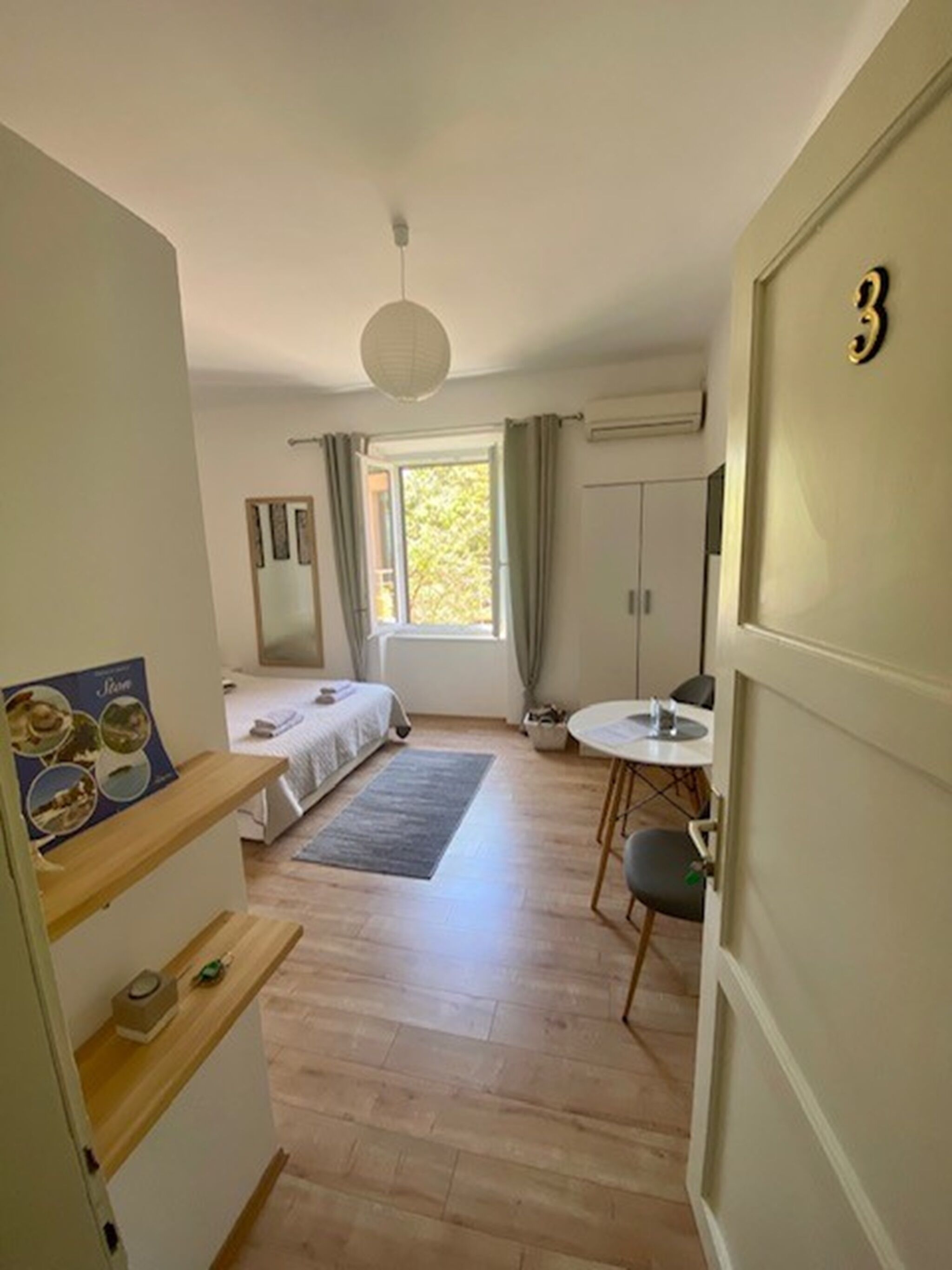 Quarto pequeno com cama, mesa e janela com cortinas.