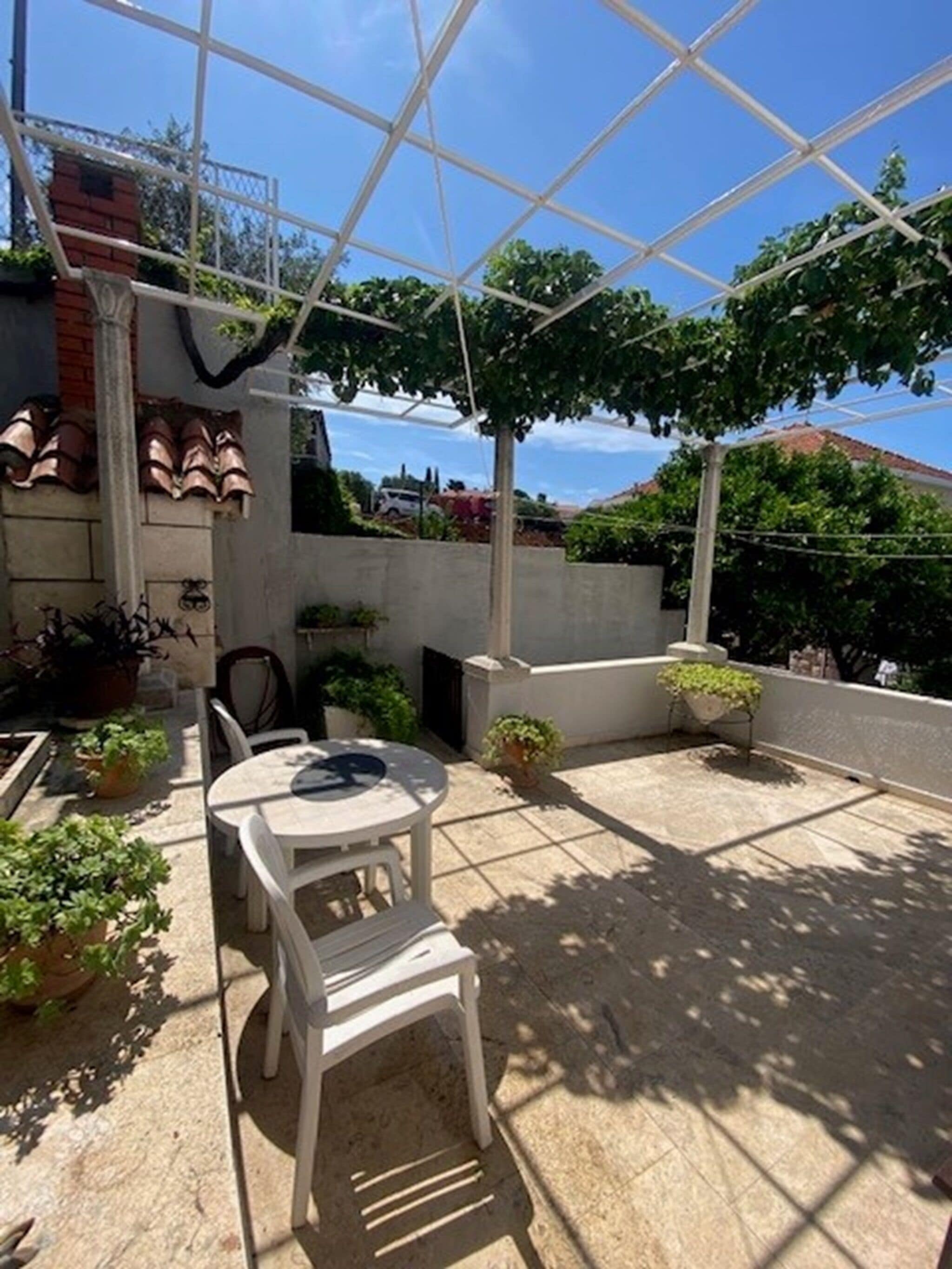 A covered outdoor patio with a table and chairs, surrounded by potted plants and a view of the neighborhood.