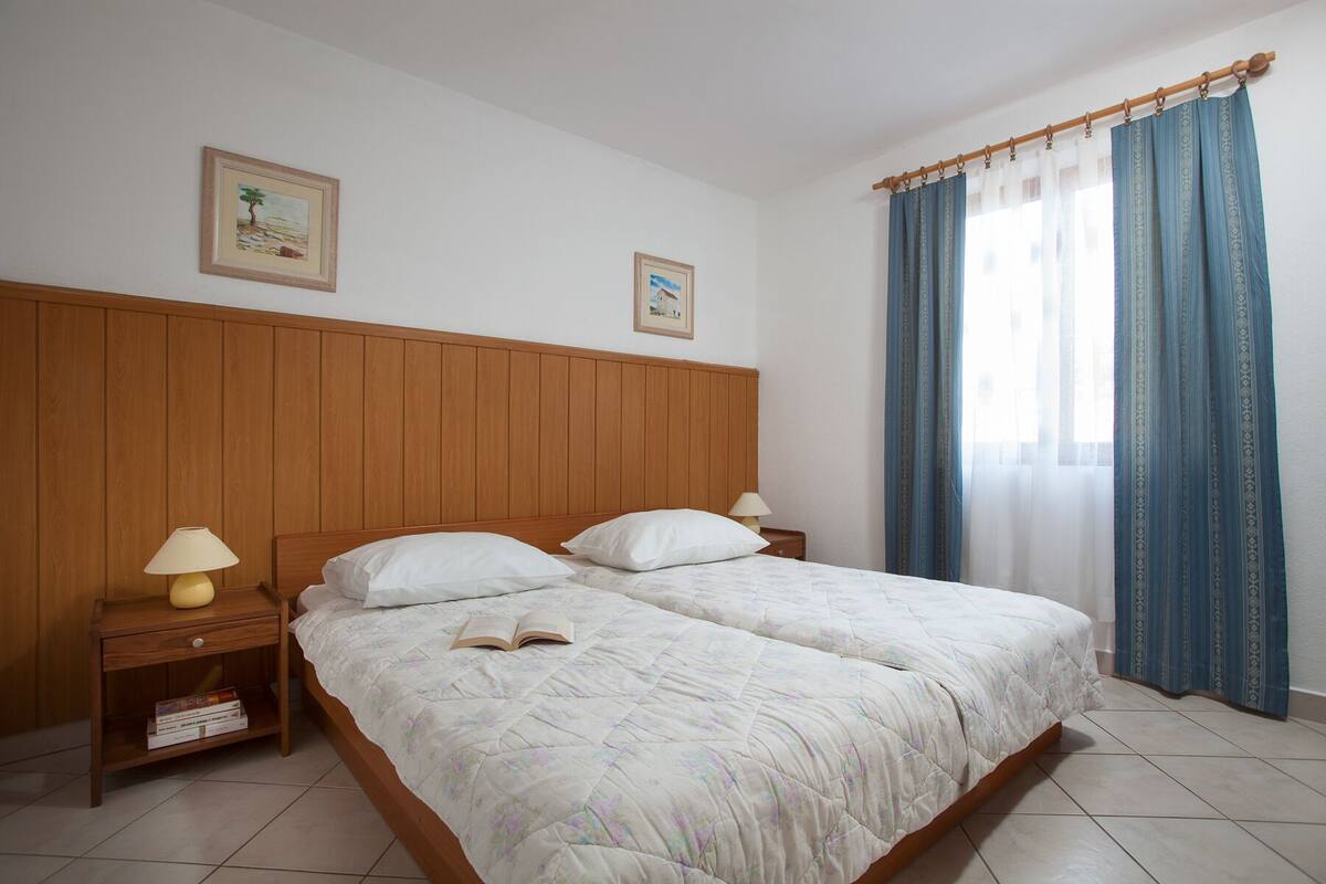 A double bed with a floral duvet, wooden headboard, two bedside tables with lamps, and a window with blue curtains.